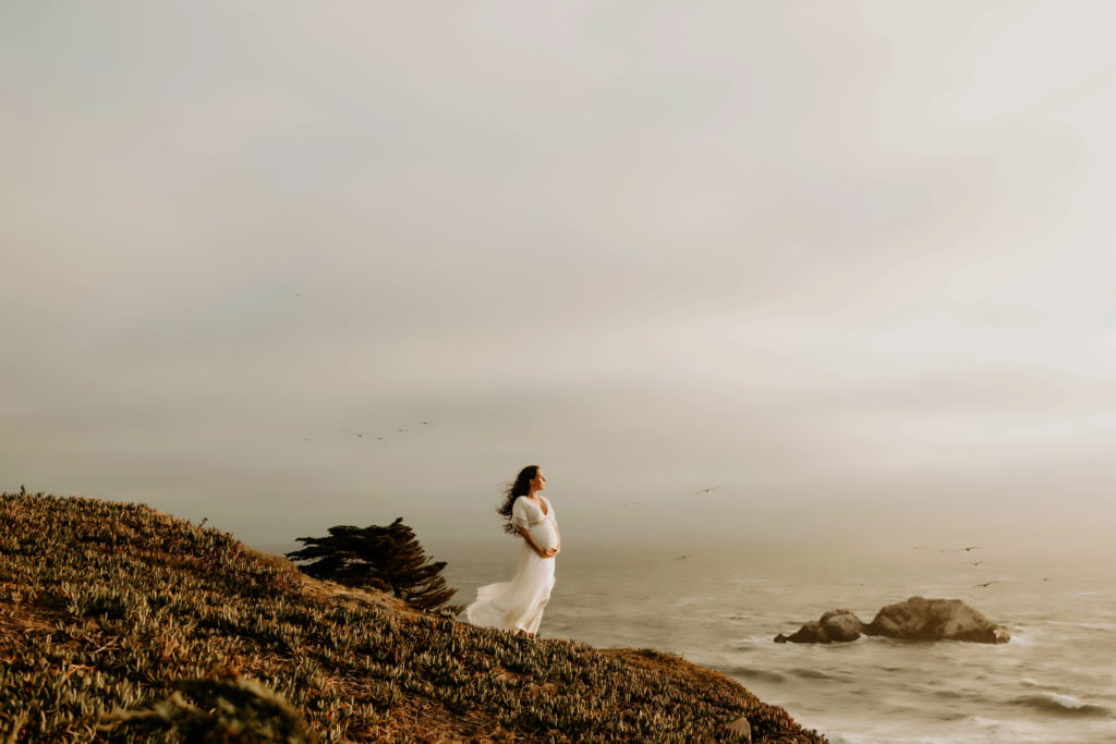 Pregnant mom-to-be standing on the edge of a mountain over looking the ocean on a cloudy day 